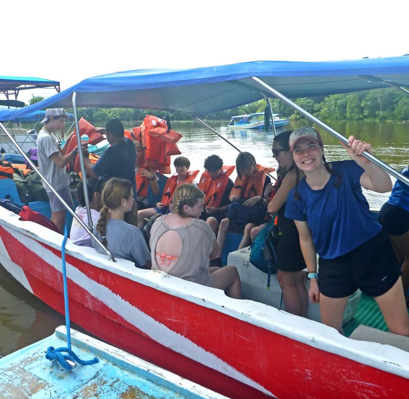 The image shows a group of people on a boat, likely tourists, wearing life jackets. The boat has a red and white striped exterior and a blue canopy. They appear to be enjoying a river or lake excursion. The background features a lush, green landscape, suggesting a tropical or subtropical environment. The overall atmosphere seems relaxed and adventurous.